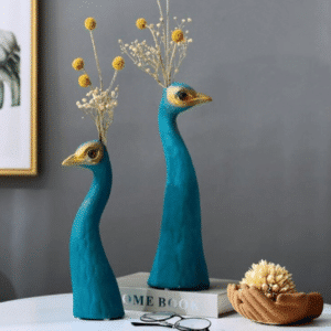 Vases à forme de tête de paon bleu exposés en deux tailles différentes. Ils contiennent des fleurs séchées jaunes et sont disposés sur une table blanche.