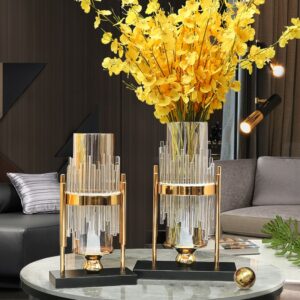 Grand vase en verre transparent en forme de tube entouré de cylindre de verre, le tout disposé sur un support en métal doré avec un socle noir. Ce vase est de style moderne, art déco, lumineux et élégant. Il contient des fleurs jaunes.