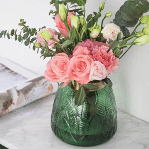 Un vase en verre à motif de feuillage sculpté. Le vase est de teinte verte, de forme ronde, avec un col doré et intègre un bouquet de fleurs composé de roses.