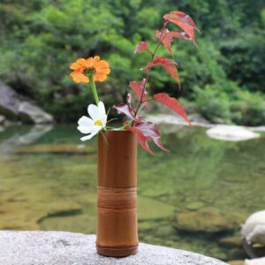 Vase en bambou creux naturel. Il est confectionné en tronc de bambou sectionné. D'influence japonaise, ce vase est naturel et minimaliste. Sa couleur est marron et il contient un bouquet de fleurs de différentes variétés. Elles sont de couleur rouge, blanche et orange. Le vase est posé sur un rocher, devant une rivière naturelle, en pleine foret.