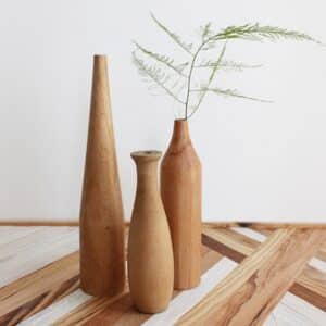 Trois vases en bois naturel en différente forme de quille. Le bois est marron clair, polis et brillant. Un des vases comporte une brindille de fougère. Ils sont exposés sur un revêtement à motif de latte de bois, devant un mur blanc.