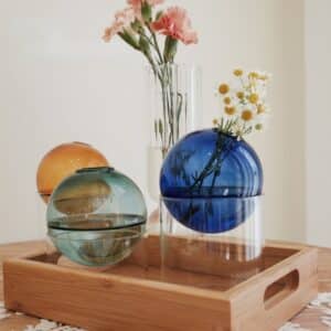 Vases en verre en forme de boules colorées. Ils rappellent la forme d'une bulle. Ils sont transparents. Un vase vert, un jaune, un bleu, et un vase tubulaire transparent en arrière-plan. Le vase bleu contient un petit bouquet de pâquerettes. Ils sont disposés sur un plateau en bois. En arrière-plan, on voit un mur blanc.