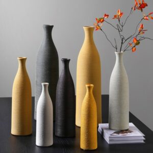 Vases en forme de bouteilles de style scandinave. Les bouteilles sont faites en céramique striée. Il y a différente taille et couleurs de vases. Petit, moyen et grand, disponible en blanc, noir ou jaune. Ils sont disposés sur une table noire. Un des vases blanc contient une branche à fleurs rouges. Le vase est surélevé par un livre blanc. En arrière-plan, on voit un mur gris.