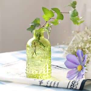 Petit vase en verre transparent de couleur vert. Il est orné de motifs géométriques gravés sur sa surface. Il contient une branche de lierre verte. Il est posé sur une table avec une nappe blanche et bleue, sur un napperon en tissus blanc et noir, à proximité d'une fleur mauve.