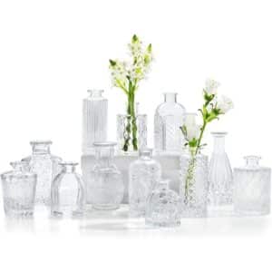 Une multitude de petits vases en verre transparent avec motifs gravés sur la surface. De tailles et de formes variées, deux vases contiennent des fleurs blanches. L'arrière-plan est intégralement blanc.