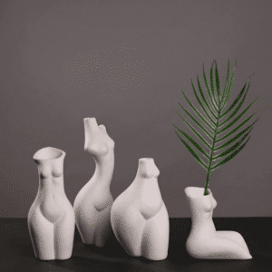 Quatre vases en forme de buste de femme nue en céramique. Les vases sont blancs, de posture et de silhouette différentes. Un des vases contient une branche de palmier verte. Les vases sont exposés sur un sol noir, avec un mur gris foncé en arrière-plan.