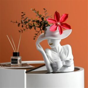 Vase en forme de buste de femme avec un chapeau. Le vase est fabriqué en céramique blanche. La femme tient son chapeau du bout des doigts. Le vase contient un mélange de fleurs séchées et d'une grosse fleur rouge. Les lèvres de la femme sont dorées. Le vase est positionné sur une sorte de meuble blanc rond, avec un diffuseur remplit de bâtons d'encens, placé sur un magazine. L'arrière-plan est un mur orange.