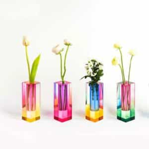 Quatre vases rectangulaires de différentes couleurs. Le premier marie le jaune et le rose. Le second, le rose et le mauve. Le troisième, le bleu et le jaune. Et le dernier, le vert et le rose. Ces vases contiennent chacun des fleurs blanches. Ils sont de style pop, avec des couleurs fluo et arc-en-ciel. L'arrière-plan et la surface sur laquelle sont disposés les vases sont blancs.