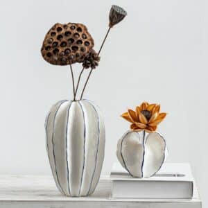 Deux vases en céramique en forme de carambole. Ils sont fabriqués en céramique. Ils sont blancs avec un liseré bleu sur les arêtes. Il y a un grand vase de forme ovale et un petit de forme ronde. Le grand vase contient des fleurs séchées de couleur marron. Le petit contient une fleur orange. Il est posé sur un livre blanc, surélevé par rapport au grand vase. Les deux sont positionnés sur une table blanche. En arrière-plan, on voit un mur gris.