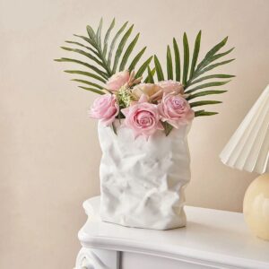 Vase blanc en céramique de forme carré. Ce vase reproduit l'aspect d'un papier froissé. C'est un trompe-l'œil. Il contient un bouquet de roses rose avec quelques feuillages verts. Il est disposé sur un meuble blanc à proximité d'une lampe beige. L'arrière-plan est un mur beige.
