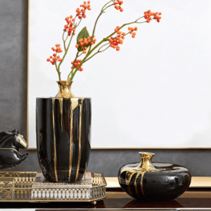 Deux vases, en céramique noire, d'aspect brillant, avec un effet de coulure dorée sur leurs surfaces. Le plus grand des deux vases contient des branches avec de petits fruits orange à ses sommités. Le deuxième vase est de forme plate et arrondie. Les vases sont entreposés sur une table avec un plateau en métal. L'arrière-plan est blanc.