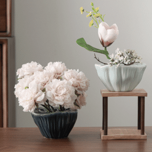 Vases en forme de coquillage, ayant la forme d'une anémone de mer. L'un est bleu foncé, posé sur une table en bois marron. Il contient des fleurs rose pâle. L'autre est bleu clair, posé sur un petit support en bois carré. Il contient une fleur rose et une petite plante à fleurs blanche. Il est surélevé par rapport au second vase. La céramique est brillante et striée. L'arrière-plan est un mur blanc-gris.