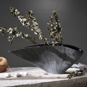 Grand vase en forme de coquillage noir. Inspiré du style japonais, il est très large et évasé. Sa céramique est brillante et finement striée. Il contient des branches de fleurs séchées. Il est entreposé sur une table en pierre avec du sable et des graviers.