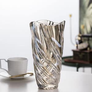 Vase en cristal strié, de forme évasé. Les striures sont diagonales. La bouche du vase est ondulée. Sa couleur est légèrement ambrée. Il est disposé sur une table, avec une tasse de café en arrière-plan.