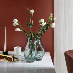 Vase en verre bleu en forme de fesses de femme. Le vase est transparent, il contient un bouquet de fleurs blanches. Il est posé sur une table blanche à côté d'une bougie en pot, d'une longue bougie, et d'un objet doré. L'arrière-plan est un mur marron avec un rideau blanc.