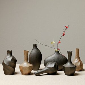 Collection de petits vases en grès minimaliste d'inspiration japonaise. Ces petits vases sont noirs ou marrons, de formes variées, abstraites ou géométriques. La céramique utilisée pour les fabriquer est brillante. L'un des vases en ovale horizontale noir contient une petite branche à fleurs jaune et rouge.