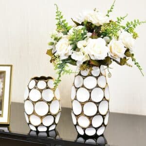 Vase moderne avec des alvéoles blanches et des interstices dorés. Il y a un vase petit format et un vase grand format. Le plus grand contient un bouquet de roses blanches avec quelques branches de fougères. Les vases sont exposés sur un meuble noir, à côté d'un cadre photo. L'arrière-plan est un mur blanc.
