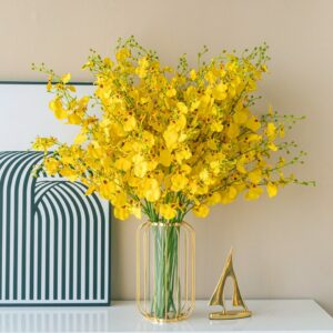 Vase moderne en forme de cage métallique dorée ovale. Le vase est constitué de barreaux dorés avec un tube de verre au centre pour contenir les fleurs. Ce vase expose un gros bouquet de fleurs jaunes. Le vase est disposé sur un meuble blanc laqué, à côté d'une sculpture de bateau dorée. En arrière-plan, on voit un tableau moderne avec des formes géométriques posé sur un mur beige.