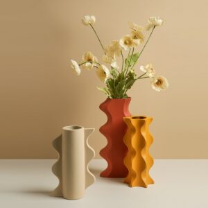 Trois vases en forme géométrique de style nordique. Un vase beige, un rouge et un orange, chacun avec des rebords ondulés, crénelés, de tailles différentes. Le vase rouge est le plus grand. Il contient un bouquet de fleurs blanches. Les vases sont exposés sur une surface blanche, avec un mur beige en arrière-plan.