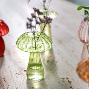 Petit vase en verre en forme de champignon. Le vase au centre est de couleur verte, il contient une brindille de fleurs violettes dans l'un des orifices qui ornent son chapeau. On aperçoit de côté un vase champignon rouge, un rose et un marron. Les vases sont posés sur une surface blanche avec des taches marron.