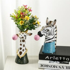 Deux vases originaux en forme de tête d'animal. Un vase est à l'effigie d'une girafe faisant une bulle avec un chewing-gum rose. Ce vase contient un bouquet de fleurs jaune et rouge. L'autre vase est à l'effigie d'un zèbre, mâchant également un chewing-gum. Ce vase est positionné sur une pile de livres. Ils sont exposés sur une surface blanche, avec un mur blanc en arrière-plan.