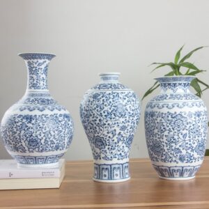 Trois vases en porcelaine blanc et bleu. Ils sont de style chinois, avec des motifs floraux et des arabesques typiques du style traditionnel. Ils ont trois formes différentes. L'un est arrondie à sa base et doté d'un col allongé avec une bouche évasée. Le deuxième à une forme d'urne, fine à la base et évasée en haut avec un col court. Le dernier à une forme ovale, plus classique, en sorte de petite jarre. Ils sont disposés sur une surface en bois marron. En arrière-plan, il y a une plante verte devant un mur gris-blanc.