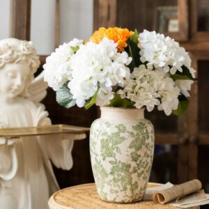 Vase en porcelaine craquelée d'aspect ancien, vintage. Le vase est blanc avec des motifs floraux de couleur verte. Il contient un bouquet de fleurs blanches et oranges. Il est posé sur une petite table en osier. On voit une statue d'ange en arrière-plan.