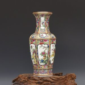 Vase en porcelaine, de forme hexagonal, de style chinois traditionnel. Ce vase en forme de jarre géométrique possède six cotés. Il est orné de motifs floraux colorés, avec du vert, du jaune, du doré, du bleu, du rose et du mauve. Il à un col allongé, une bouche évasée. Il est disposé sur un morceau de bois. L'arrière-plan est de couleur grise.