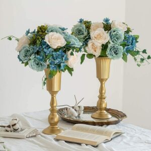Deux grands vases en forme de trompette dorée. Les vases sont en métal. Ils contiennent chacun des bouquets composés de roses bleues, roses, et d'un feuillage vert. Les vases sont disposés sur une table avec une nappe blanche, il y a un livre ouvert, un plateau en bronze. En arrière-plan, on voit un mur blanc.