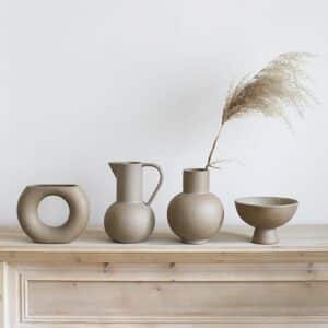 Quatre vases en terre cuite dans un style de petite poterie grecque ancienne. Ils ont chacun une forme différente : un anneau évasé, une cruche, un petit vase rond avec un col allongé et une forme de coupelle. Ils sont teintés d'un mélange de gris et de marron clair. Le petit vase avec le col contient un brin de pampa. Ils sont disposés sur un meuble en bois. En arrière-plan, on voit un mur blanc.
