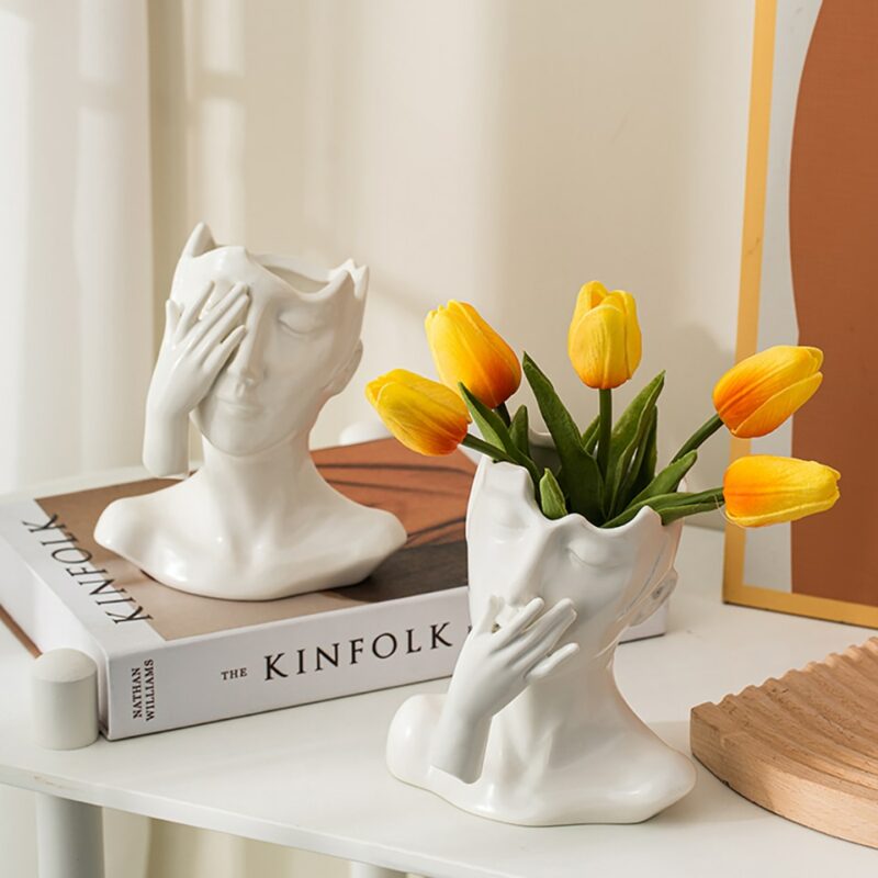 Deux vases en céramique en forme de visage de femme. L'un se cache l'œil avec la main, l'autre se cache la bouche. Ils sont tous les deux blancs. Un des vases contient des tulipes jaune orange. L'autre vase est posé sur un livre, les deux étant exposés sur une table blanche. En arrière-plan on voit un rideau blanc.