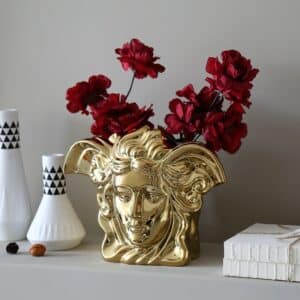 Vase doré en forme de tête de personnage mythologique grec. Il représente le visage de Médusa. Le bouquet contient des fleurs rouges. Il est disposé sur une étagère, avec d'autre petits vases blanc et noir de style art déco, et à proximité de deux livres. L'étagère et le mur en arrière-plan sont gris.