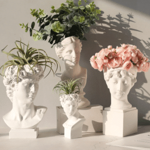 Quatre vases en forme de tête de statue grecque, mêlant des visages d'homme et de femmes. Ils sont réalisés en céramique blanche. Il y a un petit vase, et les trois autres sont en grand format. Ils contiennent chacun différentes plantes : Des plantes vertes dans trois vases et un bouquet de roses dans le quatrième. Ils sont exposés sur une surface blanche. La lumière du soleil projette leur ombre sur le mur blanc en arrière-plan.