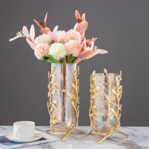 Deux jolis vases en tube de verre avec une armature en métal doré à motif de feuillage. Il y a un grand vase, qui contient un bouquet de fleurs roses et blanches. L'autre petit vase est vide. Ils sont placés sur un revêtement en marbre. Une tasse de café dans une coupelle est posée à proximité des deux vases. L'arrière blanc est gris uni.
