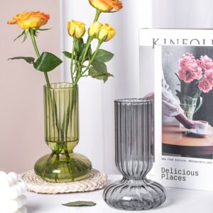 Deux vases en verre transparent en forme tubulaire avec un pied évasé et arrondi. Le verre est strié sur toute la surface du vase. Un vase est de couleur verte et l'autre grise. Le vase vert contient des roses orange. Ils sont disposés sur une surface blanche, l'un sur un dessous de plat en rotin. On voit une photo de fleurs roses en arrière-plan.