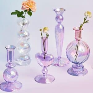 Cinq vases de forme et de couleur différente. Ils ont tous l'apparence d'un bougeoir. Un vase est couleur Lila, en forme de boule avec un col à bougie, un autre en forme de cœur couleur Lila avec un col à bougie, un autre de forme longiligne couleur Lila, un autre en forme de grosse boule couleur Lila et un dernier transparent en forme de boules superposées. Les vases sont transparents, avec des reflets arc-en-ciel et des striures sur leurs surfaces. Un des vases contient une rose orange, un autre une petite fleur jaune, et un troisième une petite fleur jaune également. Les vases sont disposés sur une surface en dégradé rose et mauve.