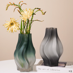 Deux vases en verre opaque. L'un est grand, de couleur verte, avec un bouquet de fleurs jaunes à l'intérieur. L'autre est soir, vide, disposé sur un livre. Les deux sont positionnés sur une surface blanche, avec un mur rose en arrière-plan.