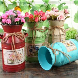 Quatre vases en forme de pot à lait ancien, de différente couleur. Ces vases sont en fer forgé. Il y a un vase rouge, un vert, un blanc et un bleu. Ils comportent des fleurs roses, blanches et rouges.