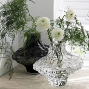 Deux vases en verre transparent de forme géométrique irrégulière. Un des vases est teinté en noir et expose un bouquet d'herbes vertes, l'autre est translucide et contient un bouquet de fleurs blanches. Ils sont disposés sur une surface beige sur fond blanc.