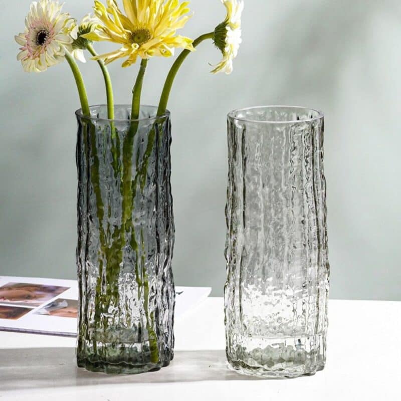 Deux vases en verre, de forme tubulaire, avec une texture irrégulière. Ces vases soufflés de manière artisanale sont de coloris variés. L'un est teinté en noir et l'autre est transparent. Le vase noir contient un bouquet de fleurs blanches et jaunes. Les deux vases sont disposés sur une table blanche, avec un mur gris en fond.