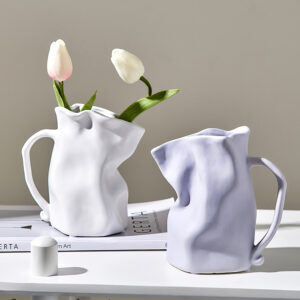 Deux vases en céramique en forme de cruche design et surréaliste. Un vase est de couleur blanche, avec deux tulipes blanche et rose à l'intérieur. L'autre vase est de couleur mauve. Ils sont disposés sur une table blanche avec un magazine en support du vase blanc. Il y a un mur gris en fond.
