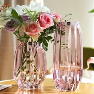 Deux vases roses en verre de forme géométrique. Les vases sont transparents. Il y a un grand vase haut et un vase plus court. Ils ont une surface anguleuse et incurvée qui leur apporte un style original. Le petit vase contient des roses de couleur rose et blanche.