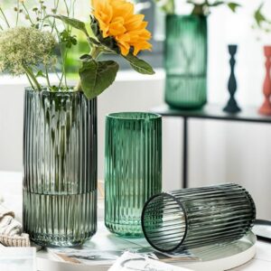 Trois vases en verre modernes, de forme tubulaire. Les vases sont striés dans le sens de la hauteur sur tout le pourtour. Il y a un grand vase gris, un petit vase vert et un autre vase gris couché. Le grand vase noir contient un bouquet de fleurs. Ils sont disposés sur une table.