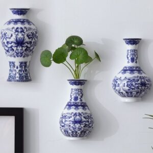 Vase suspendu bleu et blanc dans un style chinois antique. On voit trois vases accrochés sur un mur blanc, le vase central comporte des tiges de plante verte. Les vases arborent des motifs bleu et blanc traditionnels, caractéristiques du style chinois.