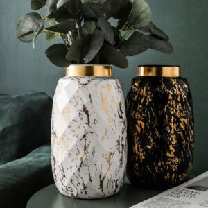 Deux grands vases modernes en forme de jarre stylisée géométrique. Les vases sont à effet marbré, l'un en noir et doré, l'autre blanc et doré. Le vase blanc contient un bouquet de plantes vertes. L'aspect du vase est texturé, sous forme d'alvéoles géométrique en petits losanges. Les vases sont exposés sur une table basse, près d'un magazine, devant un canapé. La pièce est aménagée avec du vert.