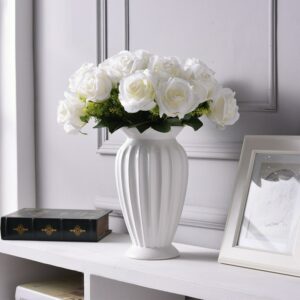 Vase céramique blanc de style classique. En forme de petit vase grec évasé et strié, c'est vase à la fois raffiné et minimaliste, dans un design épuré. Il contient un bouquet de roses blanches. Il est exposé sur un meuble blanc, à proximité d'un cadre et d'une boite en bois marron, devant un mur gris.