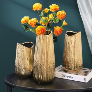 Vase moderne en céramique dorée. Le vase de forme tubulaire, légèrement évasé, est doté de striure sur son pourtour. Exposé en trois formats différents, le plus grand modèle contient un bouquet de roses orange. Les vases sont exposés sur une table noir, avec un livre en support de l'un d'eux. Le mur du fond de la pièce est vert.