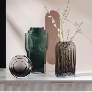 Vase moderne style art déco, de forme géométrique, teinté en gris ou vert. Un des vases contient un bouquet de fleurs blanches. Ils sont exposés sur une table blanche.