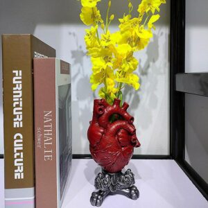 Vase coeur rouge avec un socle sculpté de couleur argent. Ce vase petit format reproduit l'organe humain à l'identique avec les valves, les artères... Il contient un bouquet de fleurs jaunes. Il est exposé sur une bibliothèque à proximité de livres.