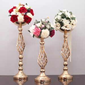 Grand vase en métal doré sculpté pour support de bouquet. On voit trois tailles de vase différentes, comportant chacun un bouquet de fleurs en forme de boule. Les vases sont exposés sur une surface marron, devant un mur gris.
